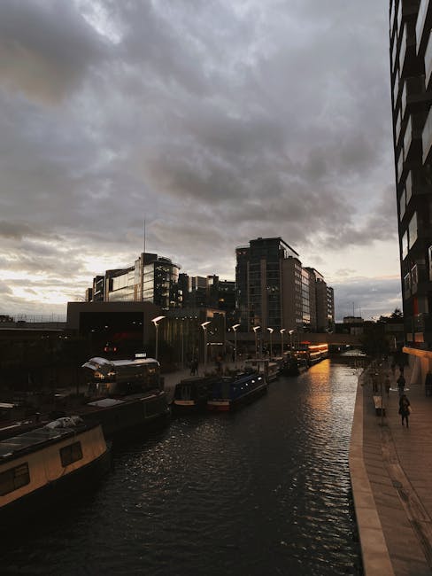 A canal scene in an urban setting during late afternoon or early evening with overcast skies. Several narrowboats and boats are moored along the water's edge on the left side, with some covered by dar