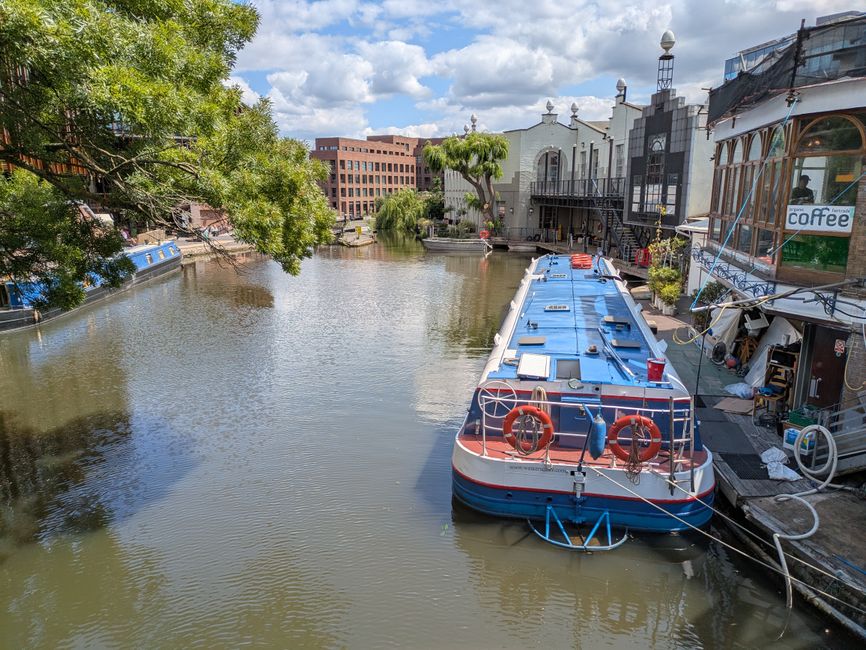 A canal scene in an urban setting during late afternoon or early evening with overcast skies. Several narrowboats and boats are moored along the water's edge on the left side, with some covered by dar